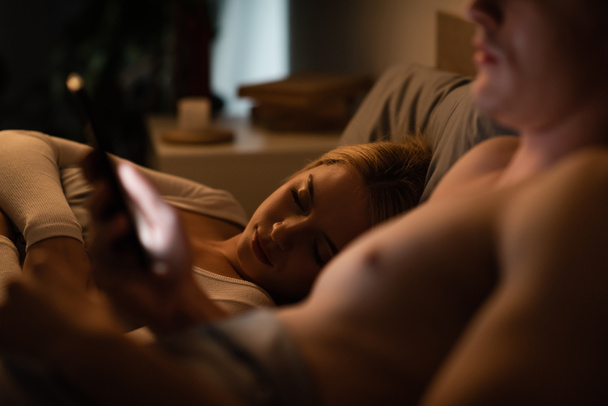 mujer rubia durmiendo al lado de novio infiel usando smartphone, concepto de engaño  - Foto, Imagen