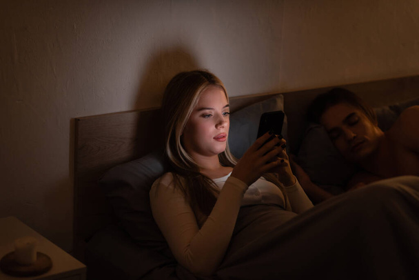 blonde woman messaging on smartphone next to sleeping boyfriend at night  - Photo, Image