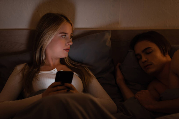 blonde woman using smartphone next to sleeping boyfriend at night, cheating concept   - Foto, afbeelding
