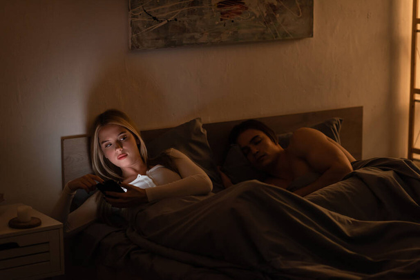unfaithful woman texting on smartphone next to sleeping boyfriend at night, cheating concept   - Foto, imagen
