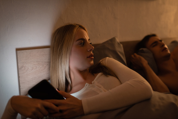 неверная женщина использует смартфон рядом со спящим парнем ночью  - Фото, изображение
