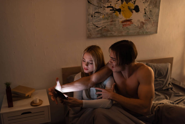angry man screaming while arguing with blonde girlfriend holding smartphone in bedroom, cheating concept  - 写真・画像