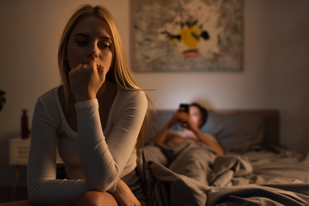 bezorgde vrouw zitten op bed in de buurt wazig vriendje met behulp van smartphone in de slaapkamer  - Foto, afbeelding