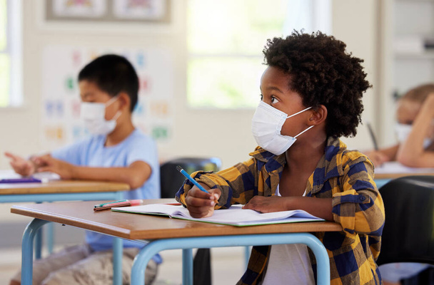 Covid, education and learning with a student in class, writing and studying at a school while wearing a mask for safety. Young boy sitting in a classroom, listening to the lesson and taking notes. - Foto, Imagen