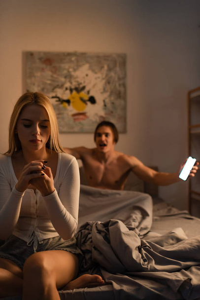 blonde woman sitting on bed next to screaming boyfriend holding smartphone in bedroom, cheating concept  - Photo, Image