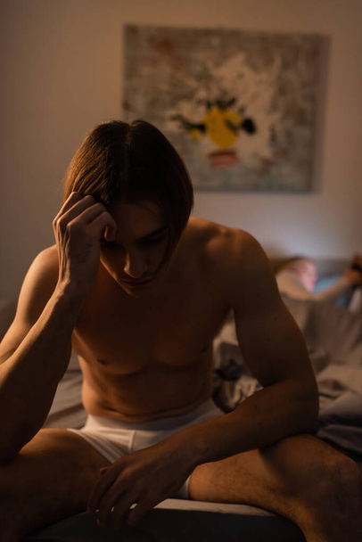 upset and shirtless man sitting on bed near blurred girlfriend, cheating concept  - Photo, Image