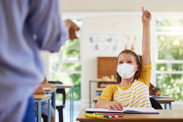 Smart student with covid face mask asking teacher question about corona virus pandemic in a classroom or elementary school. Little girl child raising hand to answer healthcare related topic in class. - Photo, image