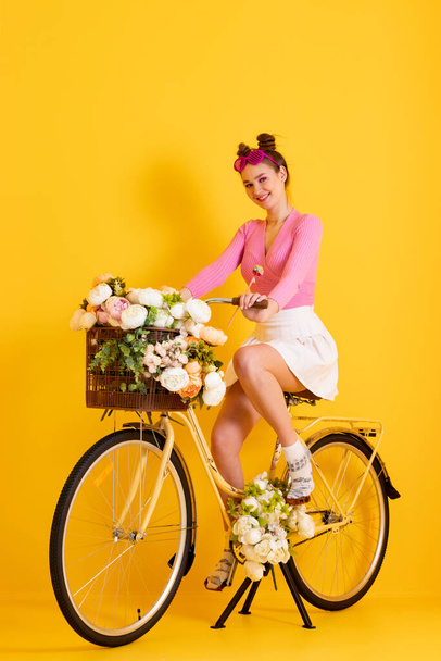 A charming, bright and happy girl in a pink top and short skirt riding a bike with a basket filled with flowers on an isolated yellow background. - Фото, зображення