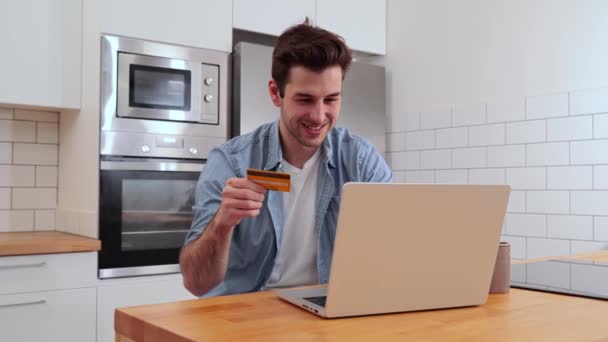 Beau caucasien jeune homme souriant et tenant une carte de crédit pour payer un achat en ligne à l'aide d'un ordinateur portable à la maison cuisine. Concept de finance bancaire. Photo de haute qualité - Séquence, vidéo