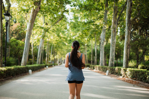 Horizontal back shot of sport young woman jogging in the park surrounded by green trees. - Фото, зображення