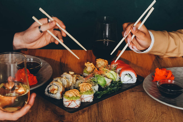 Un hombre blanco y una mujer negra comen sushi fresco delicioso y beben vino. Almuerzo en un restaurante japonés. Cocina asiática. Primer plano de las manos. - Foto, imagen