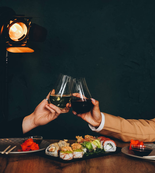 Um homem branco e uma mulher negra comem sushi fresco delicioso e bebem vinho. Almoço em um restaurante japonês. Cozinha asiática. Mãos fechadas. - Foto, Imagem