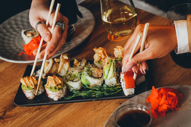 Un uomo bianco e una donna nera mangiano sushi fresco e bevono vino. Pranzo in un ristorante giapponese. Cucina asiatica. Mani in primo piano. - Foto, immagini