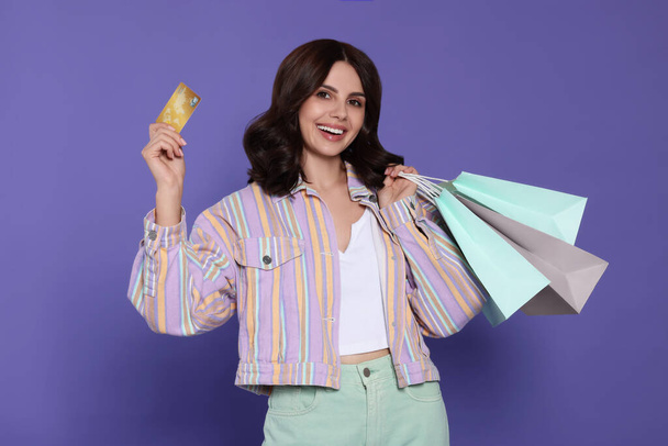 Beautiful young woman with paper shopping bags and credit card on purple background - Φωτογραφία, εικόνα