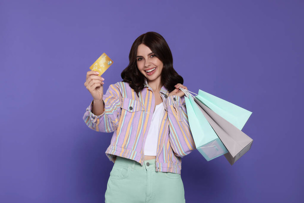 Beautiful young woman with paper shopping bags and credit card on purple background - Photo, Image