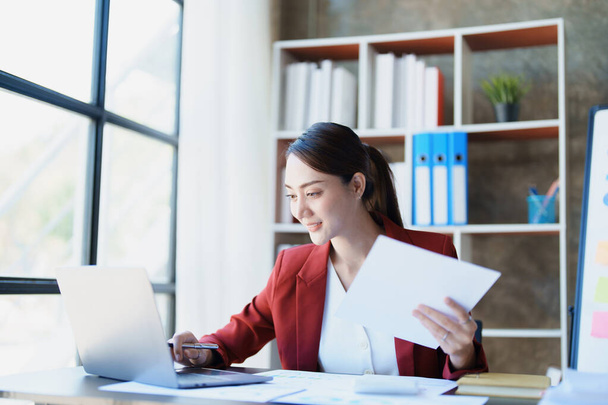 Portret przemyślanej azjatyckiej bizneswoman patrzącej na sprawozdania finansowe i przygotowującej plany marketingowe przy użyciu komputera na biurku. - Zdjęcie, obraz