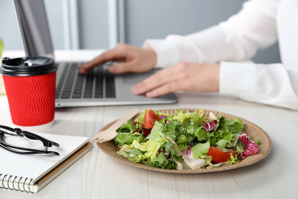 Office employee working with laptop at white wooden table, focus on vegetable salad and paper cup of coffee. Business lunch - Photo, Image