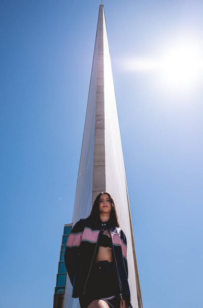 Urban chic: modelo morena joven lleva chaqueta de cuero y botas largas en gran edificio de piedra con cielo azul y luz del sol de la mañana - Foto, imagen