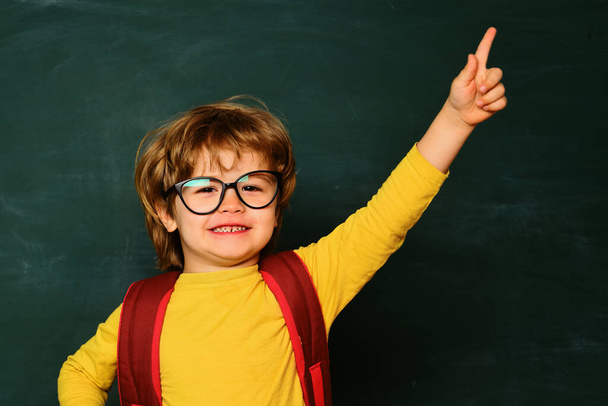 Elementary school and education. Cute boy with happy face expression near desk with school supplies. Funny little boy pointing up on blackboard - Foto, Imagen