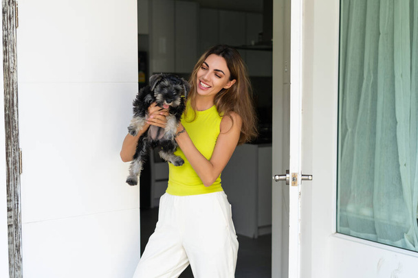 Bienvenida. Retrato de mujer alegre de pie en la puerta de un apartamento moderno, saludando al visitante y invitando a entrar en su casa, sosteniendo un cachorro - Foto, imagen
