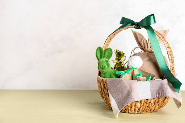Корзина с кроликом, пасхальные яйца, конфеты и подарочный пакет на зеленом столе против бежевой стены - Фото, изображение