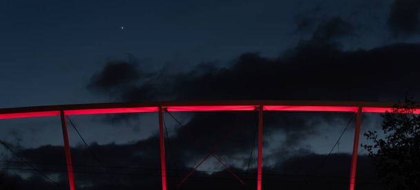 Ілюмінований фрагмент конструкції даху стадіону. Червоне світло освітлює залізобетонну конструкцію на тлі вечірнього неба
 - Фото, зображення