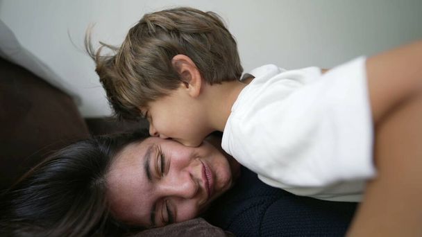 Hijo besando a madre en la mejilla. Auténtico afecto y cuidado familiar. Niño besos padre acostado en sofá - Foto, Imagen