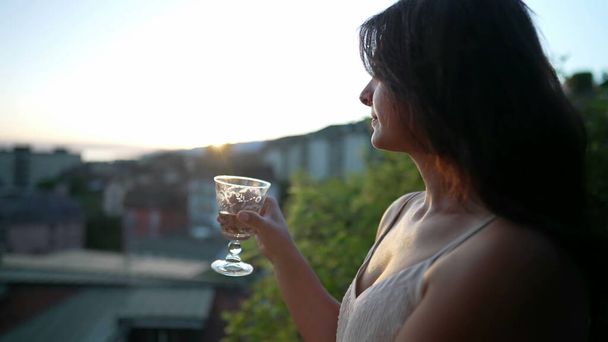 Жінка п'є біле вино на вулиці під час заходу сонця. Жінка насолоджується алкогольними напоями протягом літнього дня на відкритому повітрі, дивлячись на мальовничий вид
 - Фото, зображення