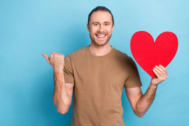 Photo of cheerful positive handsome guy dressed beige t-shirt indicating empty space hand hold heart isolated on blue color background. - Fotografie, Obrázek