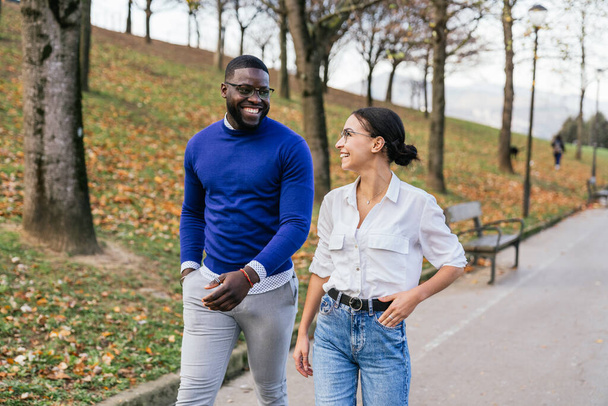 Szczęśliwy czarny chłopiec i biała dziewczyna spacerują i rozmawiają w jesiennym parku, otoczeni opadającymi liśćmi, pokazując piękno różnorodności. - Zdjęcie, obraz