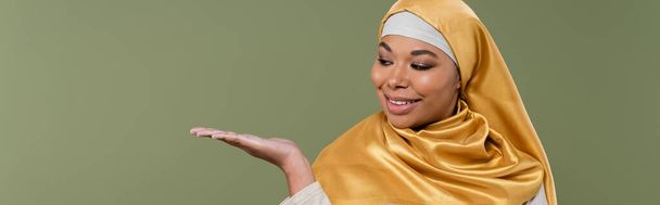 Pozytywne wielorasowa kobieta w złoty hidżab wskazując z ręki odizolowany na zielony, sztandar  - Zdjęcie, obraz