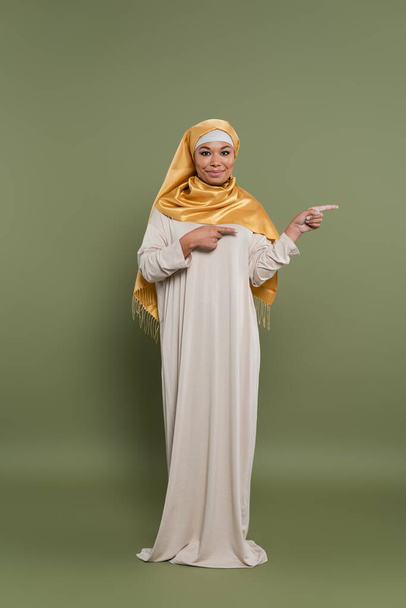 Full length of positive multiracial woman in hijab pointing with fingers on green background - Φωτογραφία, εικόνα