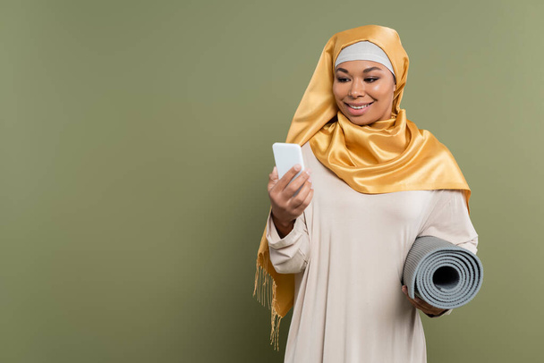 Mulher multirracial sorridente no hijab segurando tapete de fitness e usando smartphone no fundo verde - Foto, Imagem