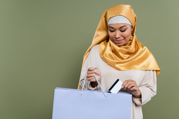 Femme multiraciale en hijab mettant la carte de crédit dans le sac à provisions isolé sur vert  - Photo, image