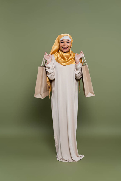 Θετική πολυφυλετική γυναίκα με μαντίλα που κρατάει τσάντες για ψώνια σε πράσινο φόντο - Φωτογραφία, εικόνα