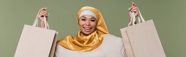 Mujer multirracial despreocupada en hijab sosteniendo bolsas de compras aisladas en verde, pancarta  - Foto, Imagen