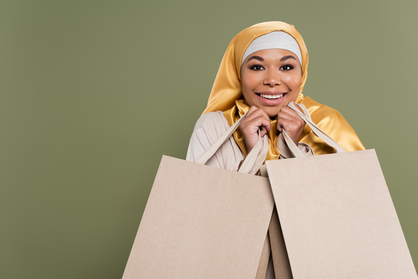 Porträt einer positiven multirassischen Frau im Hijab, die Einkaufstüten isoliert auf Grün hält  - Foto, Bild
