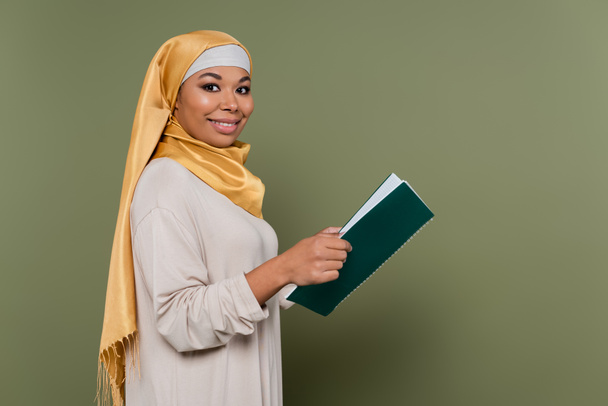 Позитивный мультикультурный студент в хиджабе держит ноутбук и смотрит в камеру на зеленом фоне - Фото, изображение