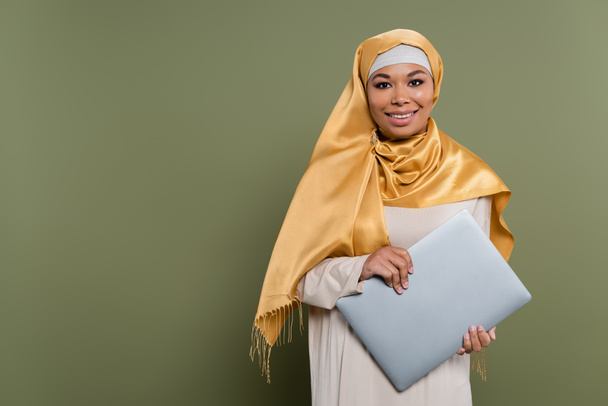 Mulher multirracial positiva no hijab segurando laptop e olhando para a câmera no fundo verde - Foto, Imagem