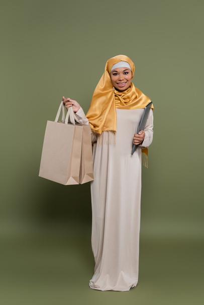 Πλήρες μήκος της πολυπολιτισμικής γυναίκας σε hijab κρατώντας φορητό υπολογιστή και τσάντες ψώνια σε πράσινο φόντο - Φωτογραφία, εικόνα