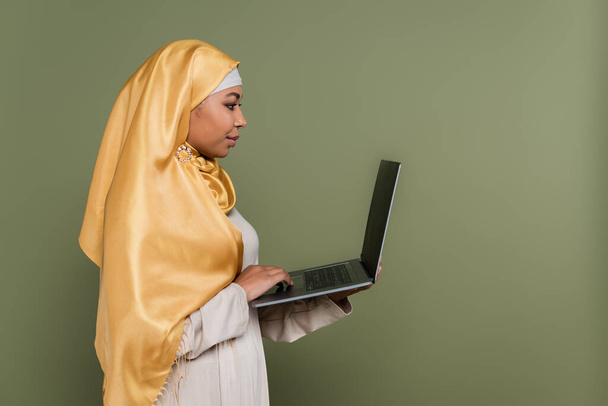 Side view of multiracial muslim freelancer using laptop isolated on green  - Fotoğraf, Görsel