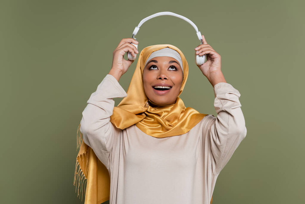 Pozytywna wielokulturowa kobieta w hidżabie trzymająca bezprzewodowe słuchawki odizolowane na zielono  - Zdjęcie, obraz