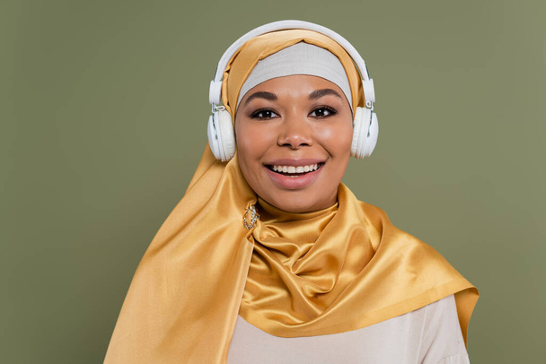 Junge multiethnische Frau im Hijab und Kopfhörer blickt isoliert auf grüne Kamera  - Foto, Bild