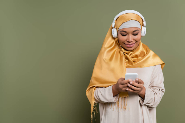 Smiling multiracial woman in hijab and headphones using smartphone on green background - Zdjęcie, obraz