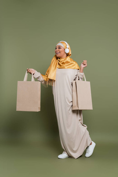 Mosolygó multiraciális nő hidzsáb és fejhallgató kezében bevásárlótáskák és séta a zöld háttér - Fotó, kép