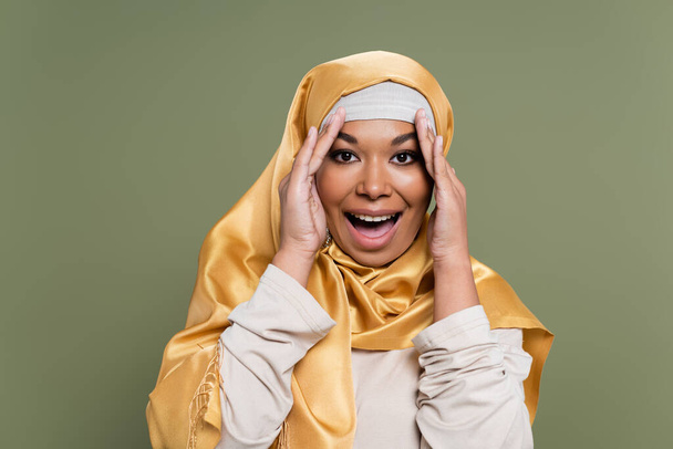 Возбужденная многорасовая женщина в желтом хиджабе смотрит на камеру, изолированную на зеленом  - Фото, изображение