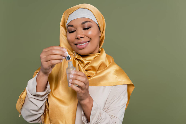 Positive multiracial woman in hijab holding cosmetic serum and dropper isolated on green  - Foto, Imagen
