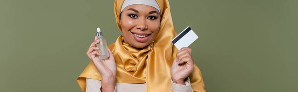 Весела багаторасова жінка в хіджабі тримає кредитну картку і сироватку ізольовано на зеленому, банер
  - Фото, зображення