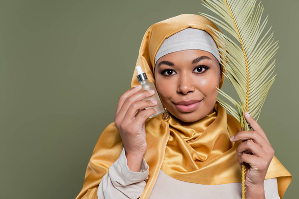 femme musulmane multiraciale en satin jaune hijab tenant feuille tropicale et bouteille de sérum cosmétique isolé sur vert - Photo, image