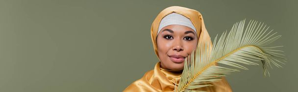 jovem mulher multirracial em hijab seda amarela sorrindo para a câmera perto da folha de planta tropical isolada em verde, bandeira - Foto, Imagem